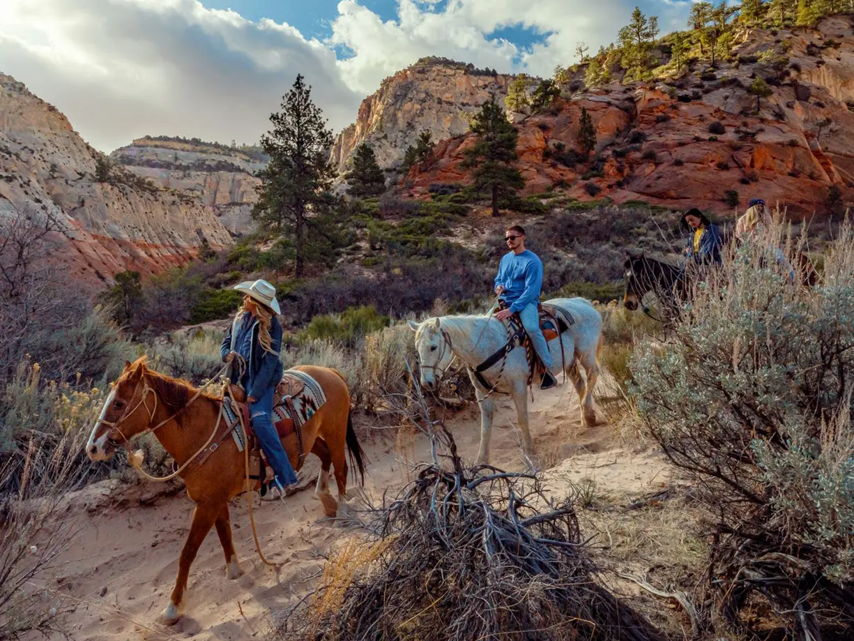 People horseback riding through a scenic desert canyon with red rock cliffs and greenery in Southern Utah.