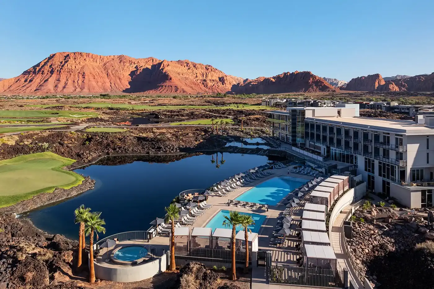 Aerial view of a modern desert resort featuring a pool and hot tub overlooking a reflective lagoon, with red rock mountains and a clear blue sky in the background.