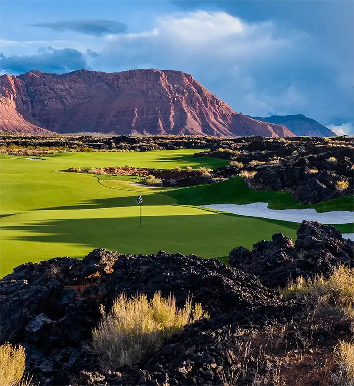 Golf course green surrounded by dark volcanic rock, with a sand bunker and flag in the foreground, set against red desert mountains under a dramatic cloudy sky.